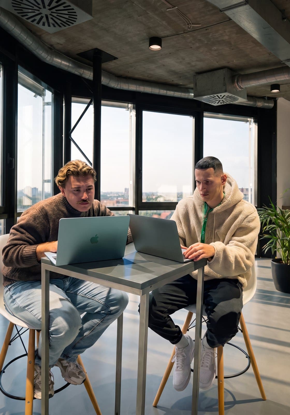 Two engineers side by side at a high table, working on two laptops in front of a window